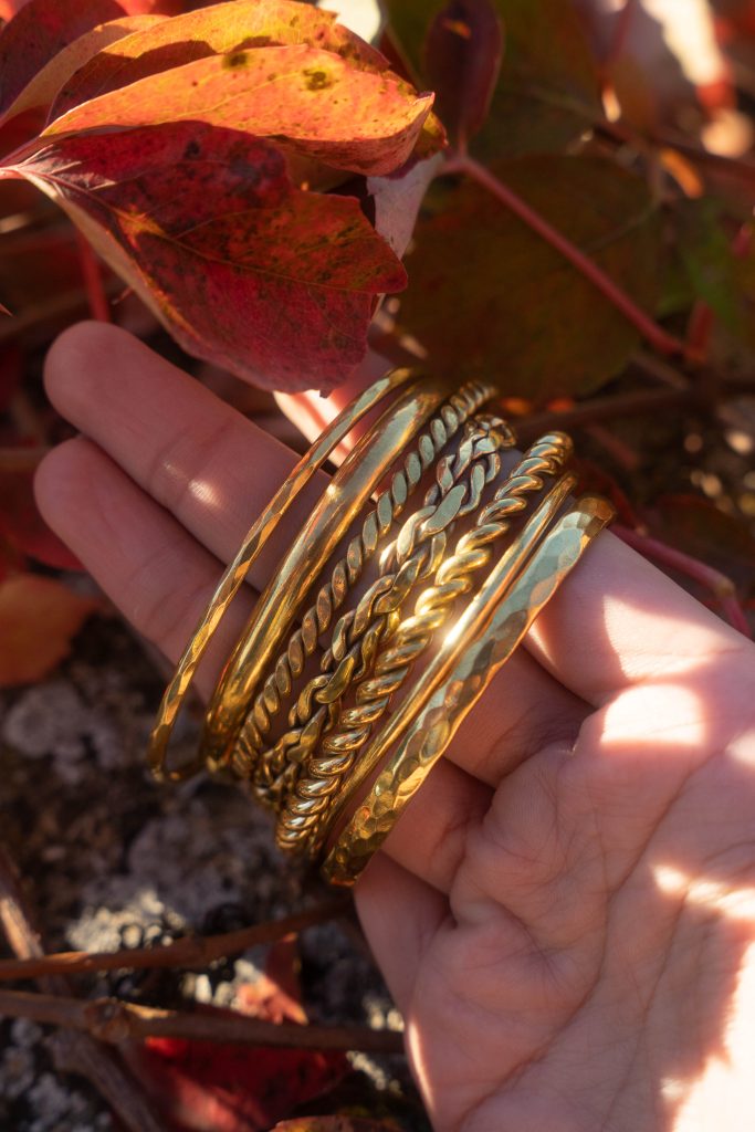 Main tenant plusieurs bracelets joncs en laiton doré (lisse, martelé et torsadé) devant des feuilles rouges d’automne, lumière chaude et naturelle.
